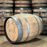 Wooden barrels with metal bands, one labeled 'GIN 270' and '12-7-23', in a warehouse setting.