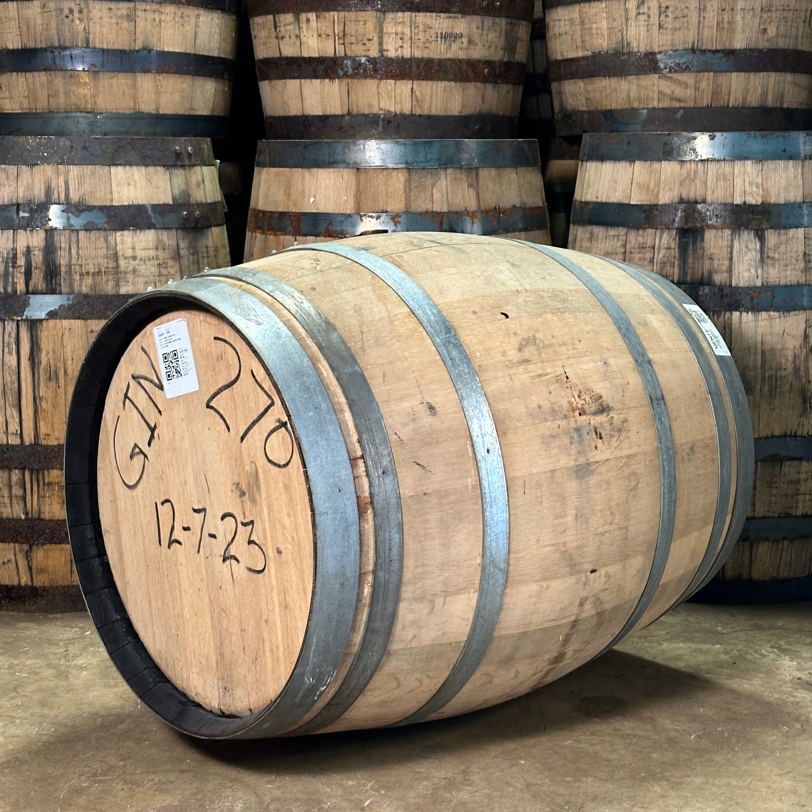 Wooden barrels with metal bands, one labeled 'GIN 270' and '12-7-23', in a warehouse setting.